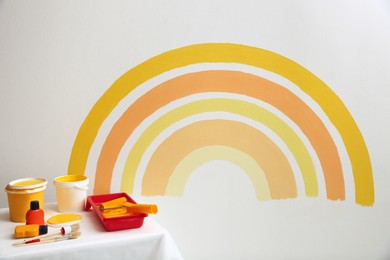 Photo of Different decorator's tools on white table near wall with painted rainbow indoors