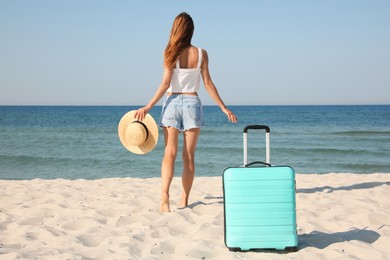 Woman walking to sea and suitcase on beach, back view