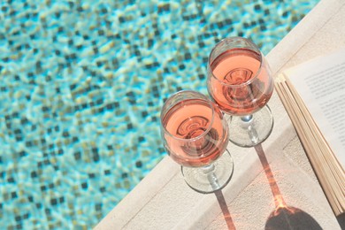 Photo of Tasty glasses of rose wine and open book on swimming pool edge, space for text
