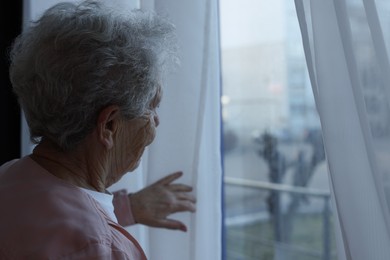 Elderly woman looking out of window indoors, space for text. Loneliness concept