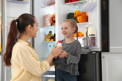 Young mother and her daughter with bottle of yoghurt near open refrigerator in kitchen