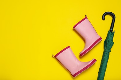 Pair of pink rubber boots near green umbrella on yellow background, flat lay. Space for text
