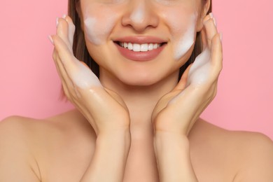 Photo of Young woman washing face with cleansing foam on pink background, closeup. Skin care cosmetic