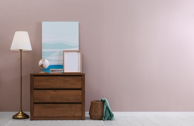 Wooden chest of drawers with globe, books and empty frame near beige wall in room, space for text. Interior design