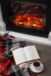 Cup of hot coffee, plaid and book on floor near fireplace, above view. Cozy atmosphere
