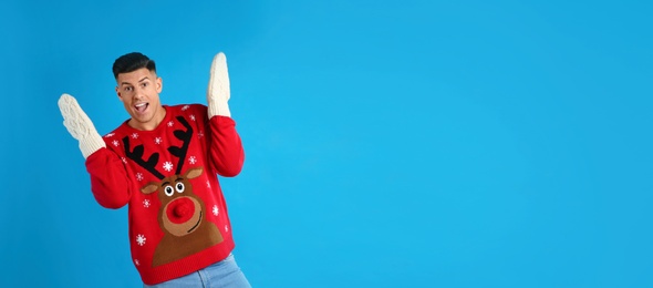 Excited man in Christmas sweater and mittens on blue background, space for text