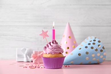Birthday cupcake with burning candle, party hats and gift boxes on pink table
