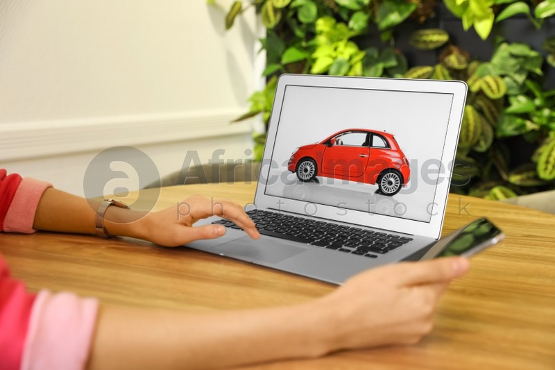 Woman using laptop and phone to buy car at wooden table indoors, closeup