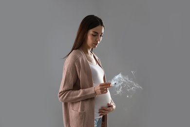 Young pregnant woman smoking cigarette on grey background
