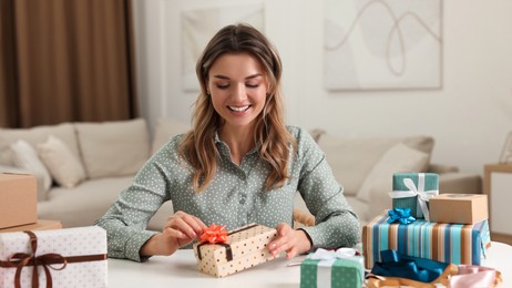 Beautiful young woman decorating gift box with bow at table in living room