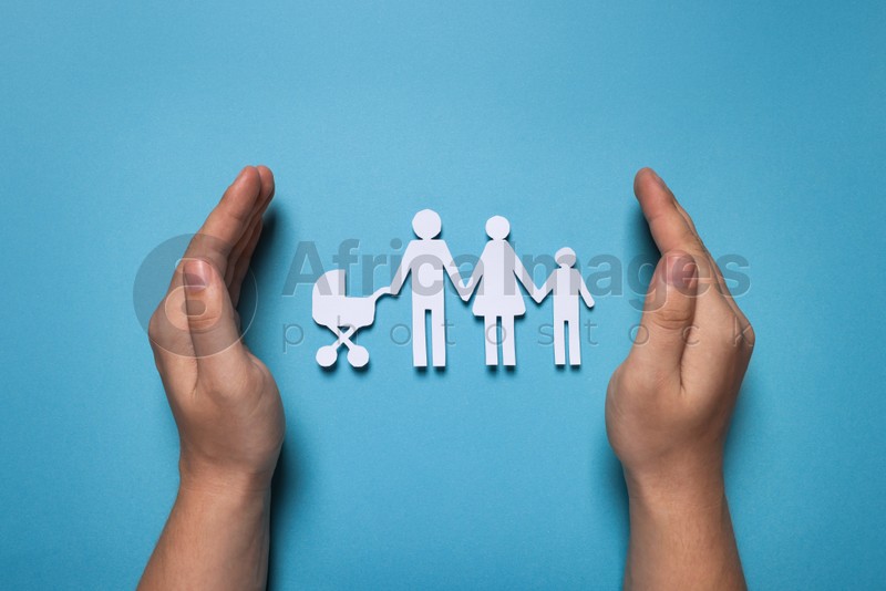 Photo of Man protecting paper family figures on light blue background, top view. Insurance concept