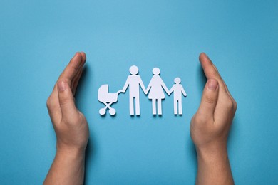 Man protecting paper family figures on light blue background, top view. Insurance concept