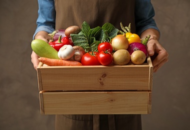 Man holding wooden crate full of fresh vegetables on brown background, closeup