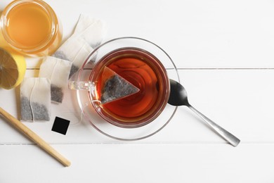 Tea bags, honey and lemon near cup of hot drink on white wooden table, flat lay