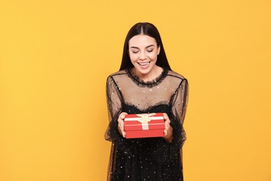 Happy young woman in festive dress holding gift box on orange background