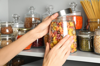 Woman taking jar of colorful pasta from white shelf, closeup