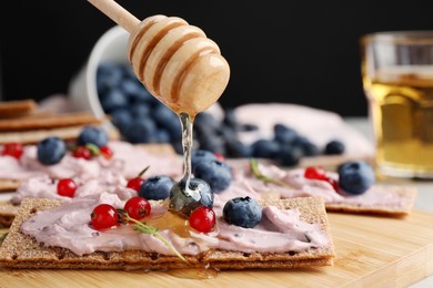 Pouring honey onto tasty cracker sandwich with cream cheese, blueberries, red currants and thyme on wooden board, closeup