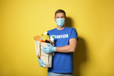 Male volunteer in protective mask and gloves with products on yellow background. Aid during coronavirus quarantine