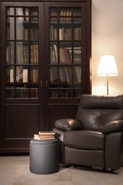 Comfortable armchair with pouf, books and lamp near wooden bookcase in library