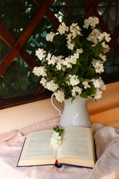Bouquet of beautiful jasmine flowers in vase and open book on fabric indoors
