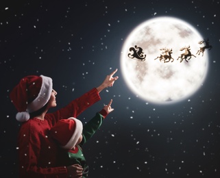 Mother and her little daughter looking at Santa Claus with reindeers in sky on full moon night. Christmas holiday