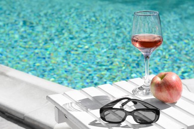 Photo of Glass of tasty rose wine, sunglasses and apple on wooden table near swimming pool, space for text