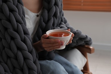 Woman with chunky knit blanket and cup of tea in armchair at home, closeup