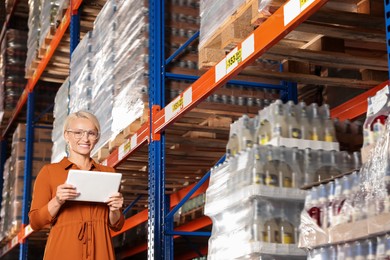 Photo of Happy manager holding modern tablet in warehouse with lots of products, low angle view