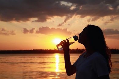 Young woman drinking water to prevent heat stroke near river at sunset, space for text