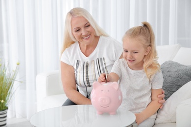 Little girl putting money into piggy bank and her grandmother at table