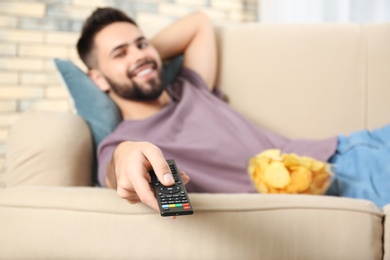 Young man with remote control watching TV on sofa at home, focus on hand