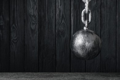 Prisoner ball with chain hanging near black wooden wall, space for text
