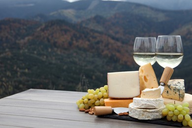 Different types of delicious cheeses, snacks and wine on wooden table against mountain landscape. Space for text