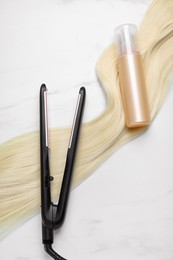 Photo of Spray bottle with thermal protection, lock of blonde hair and modern straightener on white marble table, flat lay
