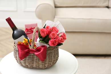 Wicker basket with gift, bouquet and wine on white table indoors. Space for text