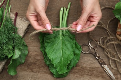 Woman tying bunch of fresh green leaves with twine at wooden table, top view. Drying herbs