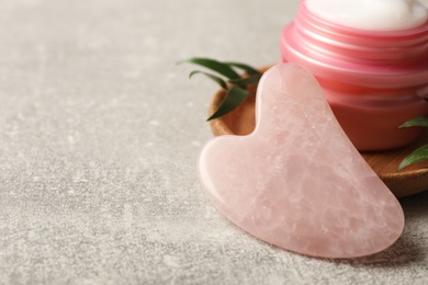 Photo of Rose quartz gua sha tool and jar of cream on grey table, closeup. Space for text
