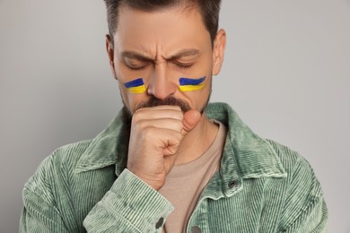 Photo of Sad man with drawings of Ukrainian flag on face against light grey background, closeup