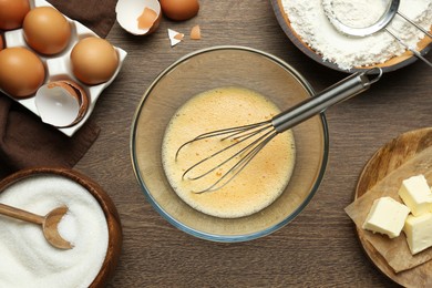 Beaten eggs, whisk and ingredients on wooden table, flat lay