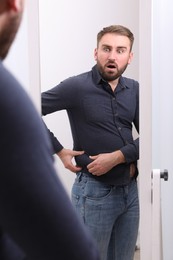 Scared man wearing tight shirt in front of mirror at home. Overweight problem