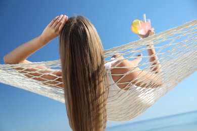 Young woman with refreshing cocktail relaxing in hammock on beach