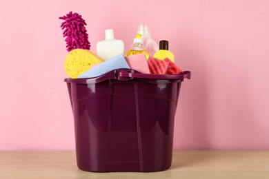 Bucket with different cleaning supplies on wooden floor near pink wall