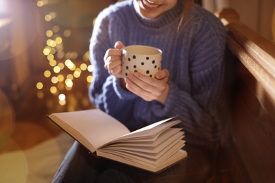 Woman with cup of hot beverage reading book at home in winter evening, closeup
