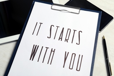 Clipboard with phrase It Starts With You, tablet and smartphone on white table, closeup