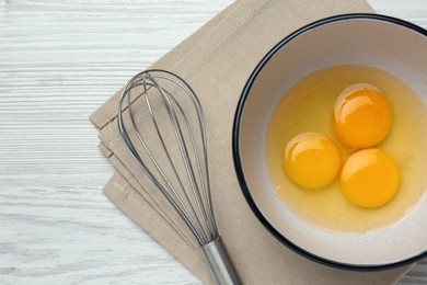 Whisk near bowl with eggs on white wooden table, top view. Space for text