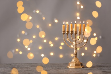 Golden menorah with burning candles on table against grey background and blurred festive lights, space for text