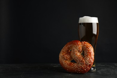 Tasty pretzel and glass of beer on black table against dark background, space for text