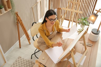 Young woman drawing on easel with pencil at table indoors, above view