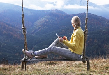 Young woman drawing with graphic tablet while sitting on swing in mountains