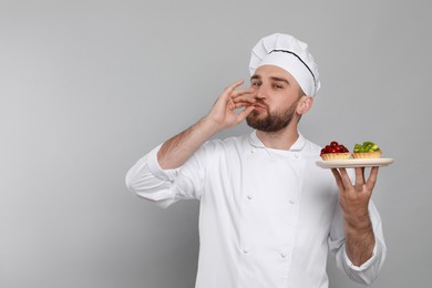 Photo of Happy professional confectioner in uniform with tartlets showing delicious gesture on light grey background. Space for text
